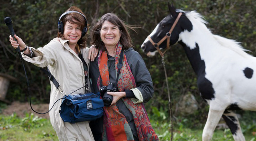 The two Kitchen Sisters stand close smiling with a white and black horse.