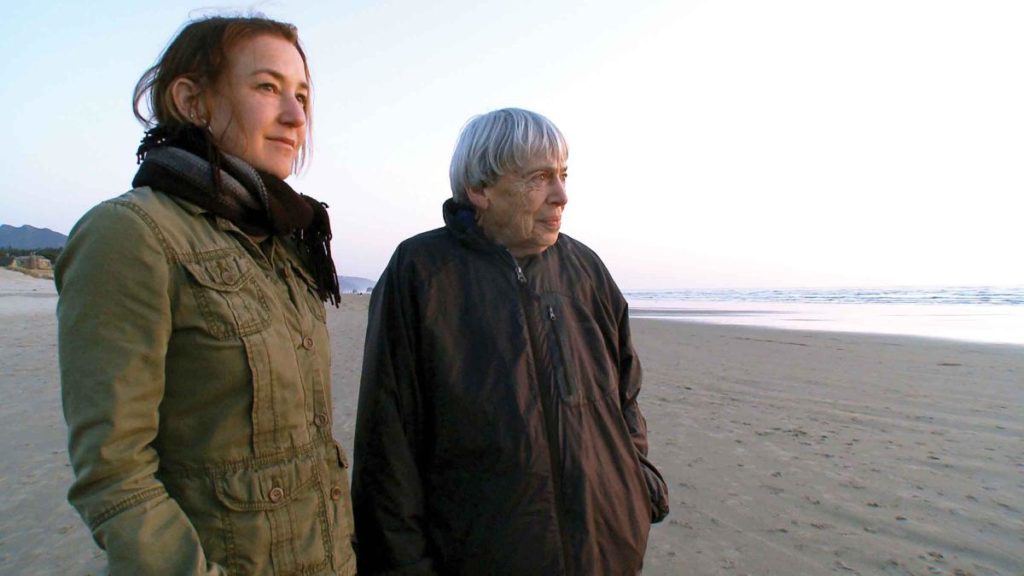 Two people stand next to each other on a beach. It is dusk and the light is dim. Both wear sturdy jackets, and the one closer to the camera has long hair pulled back and a scarf. Some of their hair is loose and flying in the wind. The other standing further back looks older and has gray hair in a circular cut. They both look at something off in the distance.