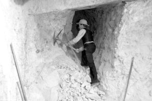 A black-and-white photo shows a man wearing a hat with a pick and work clothes inside of a rock formation, picking away the rubble. 