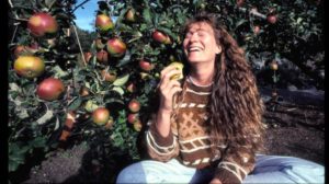 A woman sits under an apple tree holding an apple with a bite taken out of it. She is laughing.
