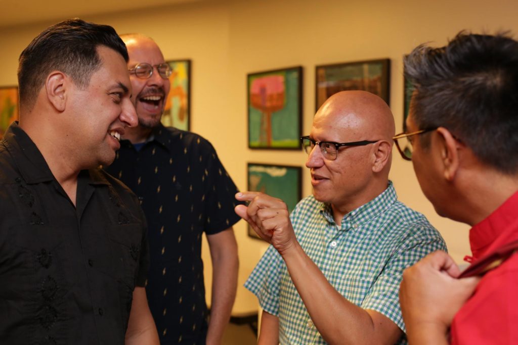 A man wears glasses, speaking, and gesturing in the middle of a group of other men who are laughing, smiling, and listening attentively.