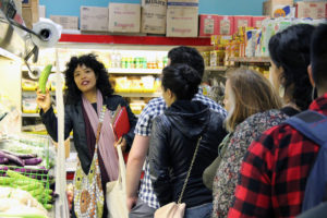 A woman speaks to a small group of listeners as she holds up a bitter melon inside a grocery store. 