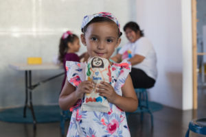 A young girl holds a small piece of artworks he made. Behind her are more children and parents.