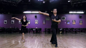 A man and woman are in a ballroom with wooden floors. They both wear all-black outfits, and stand apart, performing the same dance moves.
