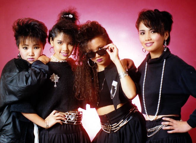 A group of four young Asian girls pose together wearing 1980s fashion against a pink backdrop.