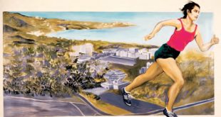 A woman, who is wearing a pink tank top and black running shorts, running up a hill with a city and the ocean below her.