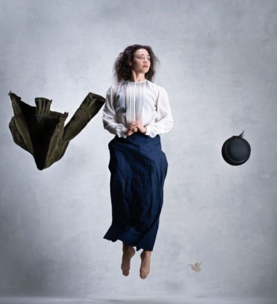 Person jumping straight in the air with long brown hair, white blouse, and blue skirt. A jacket and hat float on both sides of their body.