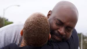 A person with brown skin and a bald head grins with their eyes closed as they hug another person with short light brown hair.