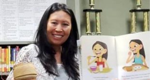 A person with long dark hair holding a children's book with colorful illustrations in one hand and a small rice pot in the other hand. Behind her are shelves of books in a library.