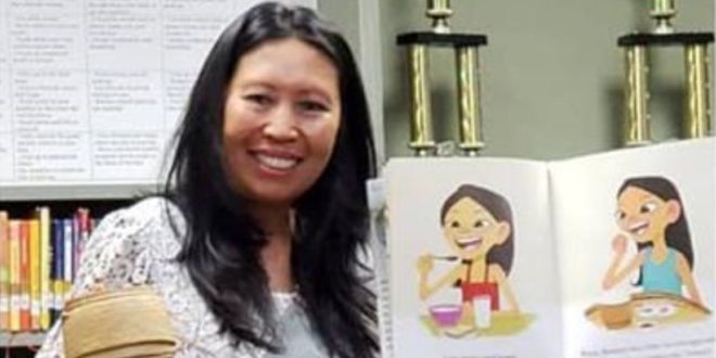 A person with long dark hair holding a children's book with colorful illustrations in one hand and a small rice pot in the other hand. Behind her are shelves of books in a library.