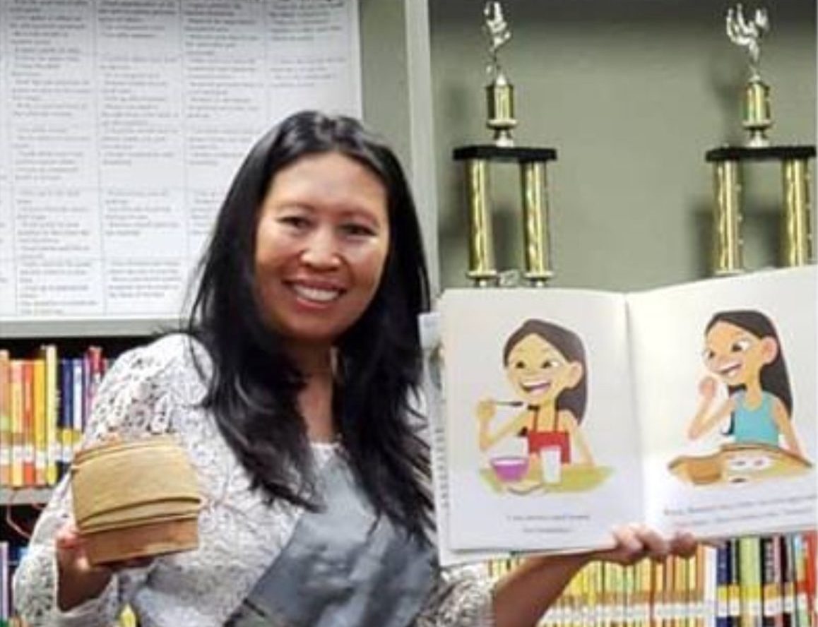 A person with long dark hair holding a children's book with colorful illustrations in one hand and a small rice pot in the other hand. Behind her are shelves of books in a library.