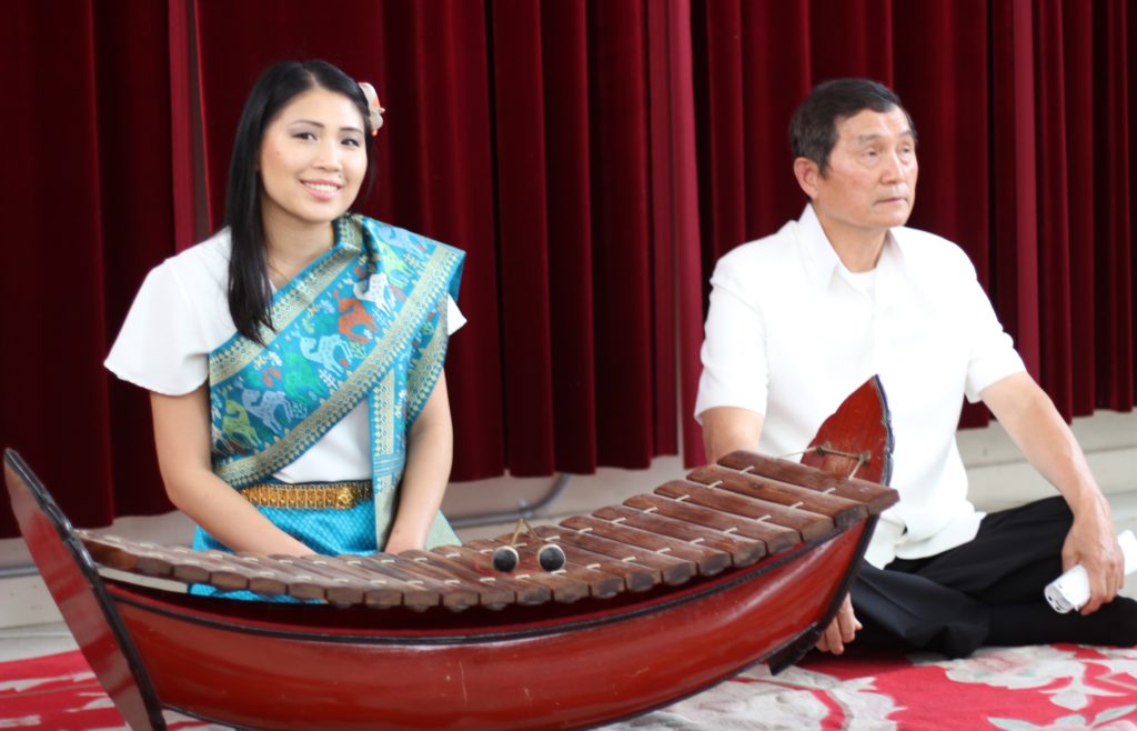 Sherina Han Khampraseuth - a master performer in lanard (or lanat), sits with another person behind a suspended, wooden xylophone.