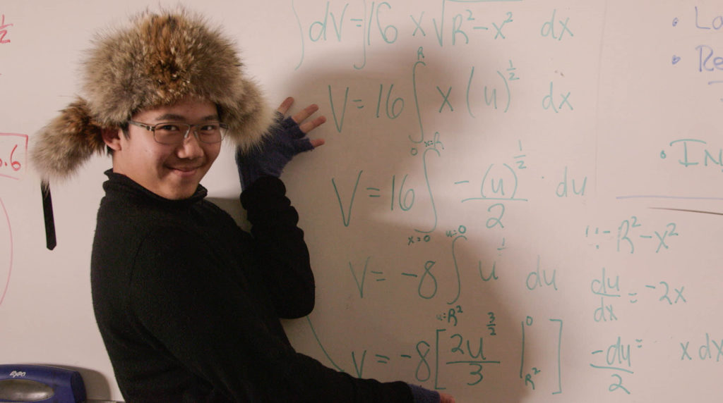 A teenage student sits next to a whiteboard with math equations on it.