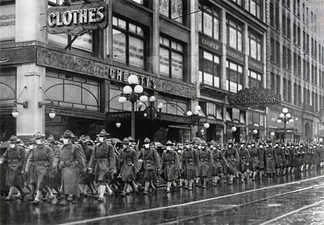 Rows of soldiers march through a city street with tall buildings.