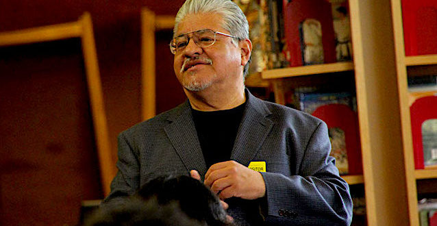 A photo of Luis Rodriguez speaking in a library wearing a grey and black suit. 