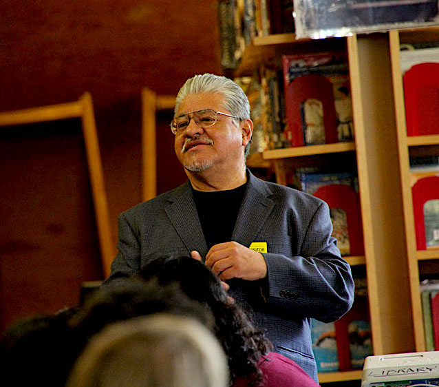 A photo of Luis Rodriguez speaking in a library wearing a grey and black suit. 