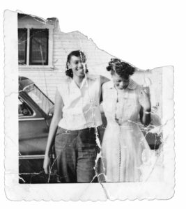 Two persons stand next to an older car with their arms connected. The photo is black and white and ripped on the top corner.