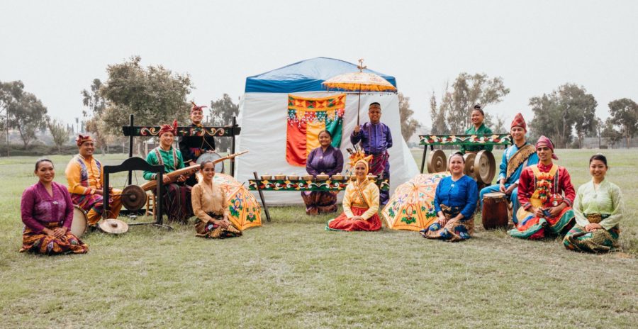 A large group of persons sit on the grass facing the camera. They are dressed in colorful and traditional clothes. Many types of instruments surround them.
