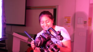 A youth holds a video camera and smiles while looking at it.
