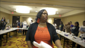 A black mother stands in front of long tables with people standing behind them holding papers.