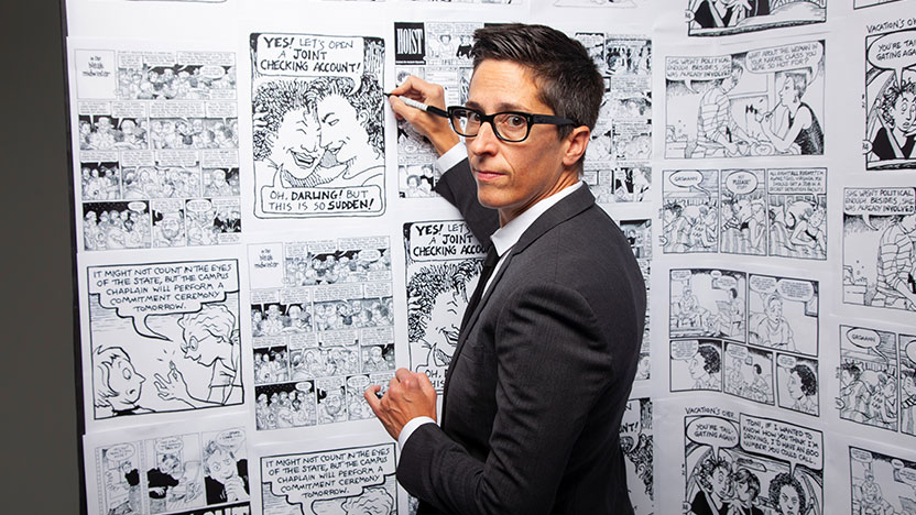 A person wearing a black suit holding a pen stands in front of a large wall of black and white comic illustrations.