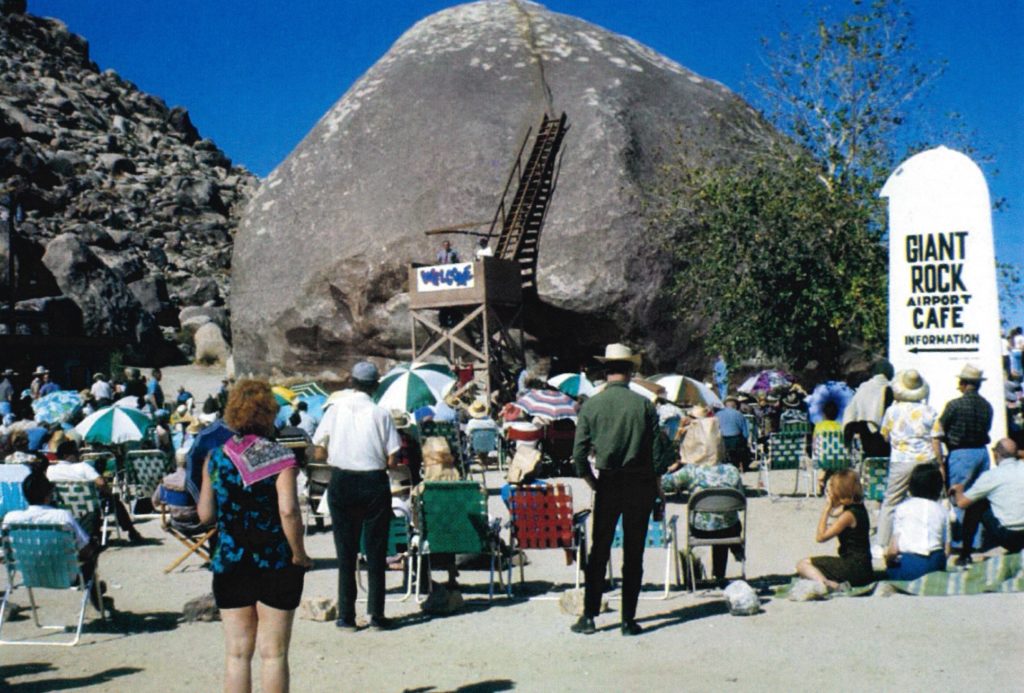 A large group of people stand and sit in lawn chairs facing the Giant Rock.