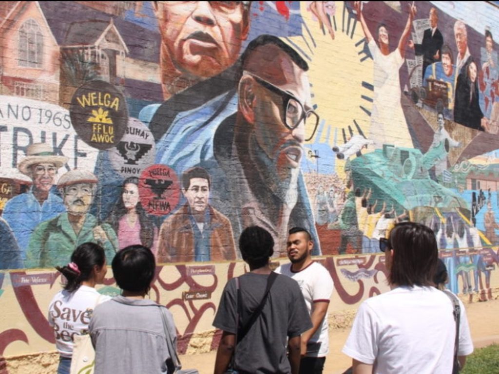 A group of persons stand in front of a building-size mural.