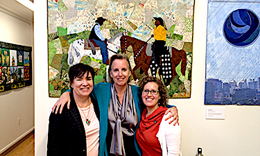 A group of three persons stand close together in front of a hanging quilt.
