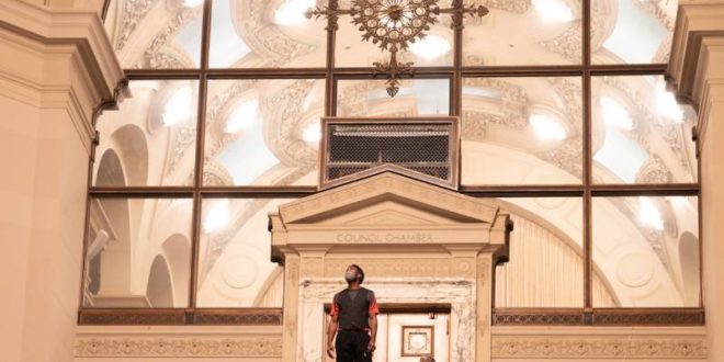 A performer stands in the center of a large staircase in front of a giant backdrop inside city hall.