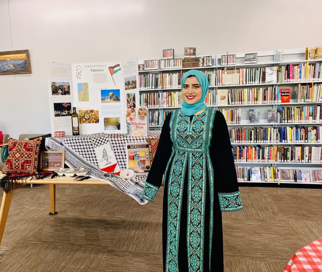 A person dressed in a Palestinian embroidered green dress with a headscarf stands in front of a display featuring Palestinian culture items.