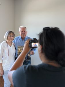 A couple holds a photo as they look at a person taking their picture.