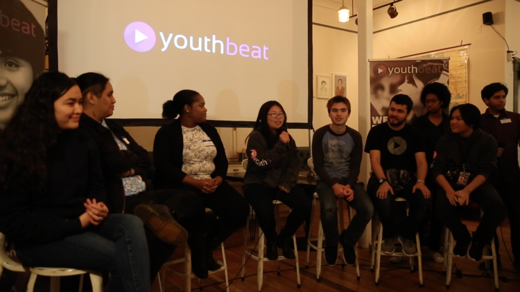 A panel of speakers sit on stools. A screen that says youth beat is behind them.