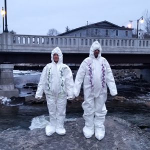 Two people wearing white bulky hazmat suits stand outside in front of a bridge.