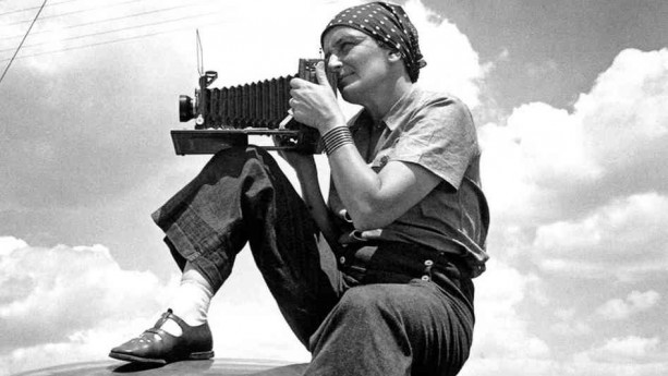 Black and white photo of. woman wearing a bandanna on her head and pants holding a camera.