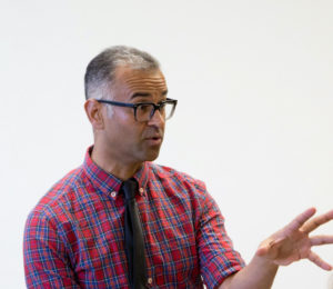 A man wearing glasses and a plaid shirt with a tie is speaking with his hand outstretched.