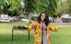 An African American woman stands in a park speaking into a microphone. She is has long black hair and is wearing a colorful robe and striped dress.