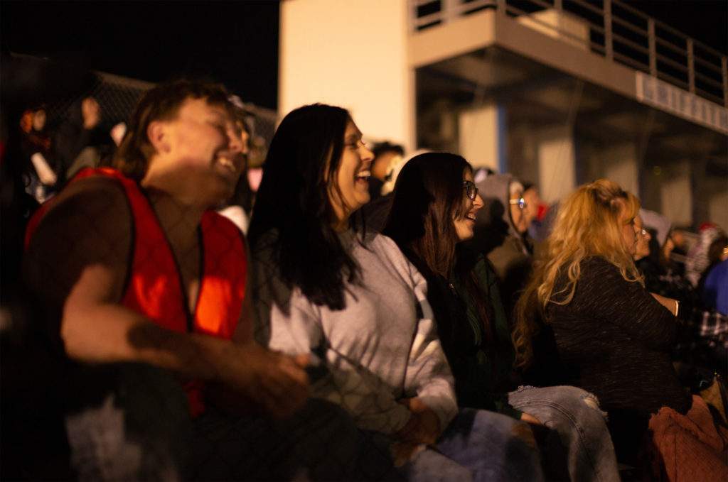 A group of people sitting outside on bleachers at night are laughing