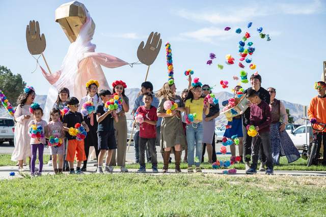 A group of children with colorful flowers are standing outside in front of a large puppet.