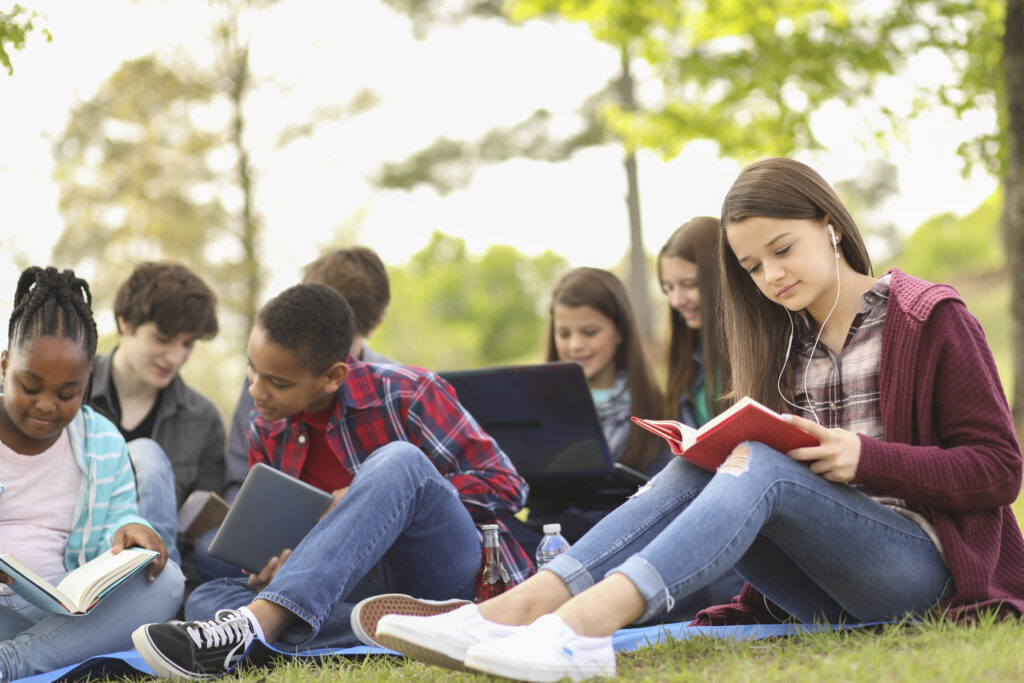 Middle grade youth sitting on the grass and reading, trees in the background