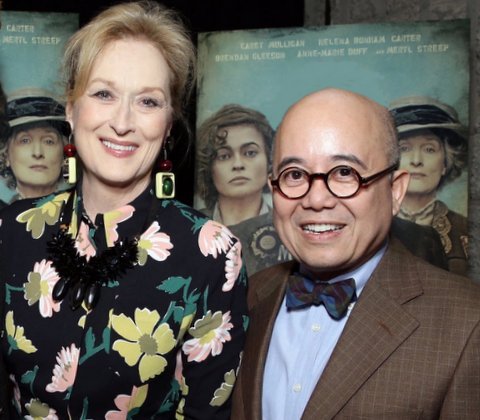 A woman with a flowered blouse and a bald man with glasses smile for the camera in front of film posters.
