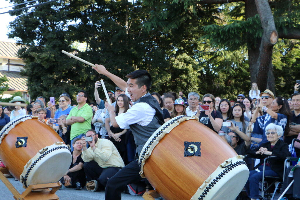 Boy enthusiastically bangs a taiko drum in front of a crowd on a street.
