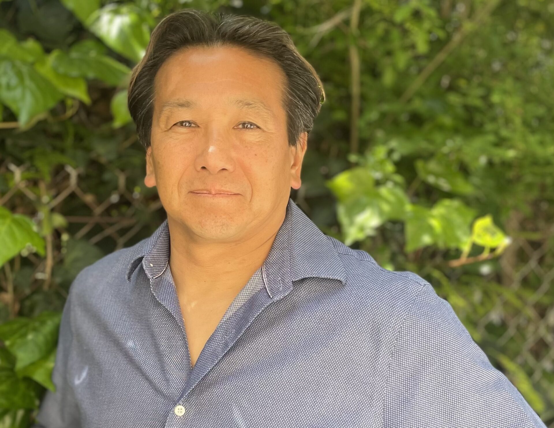 A man wearing a blue button-up shirt posing for the camera in front of trees, hands on his hips.
