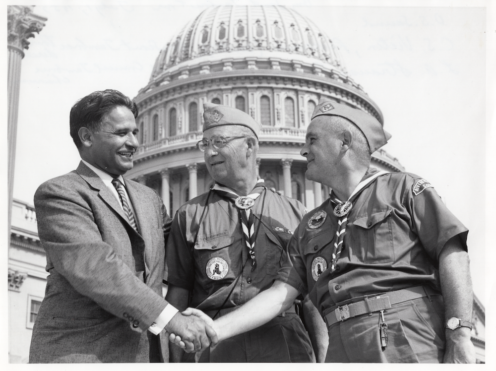 Black and white photo of man shaking hands with another man wearing a scout uniform. US Capitol in the background.