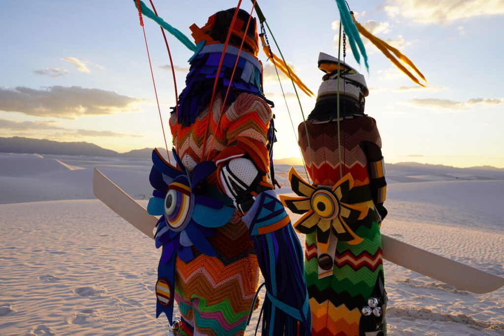 A still from Winyan Yamni / Three Dreams, showing two figures wearing colorful garments with backs to camera standing on white sand and looking at the sunset