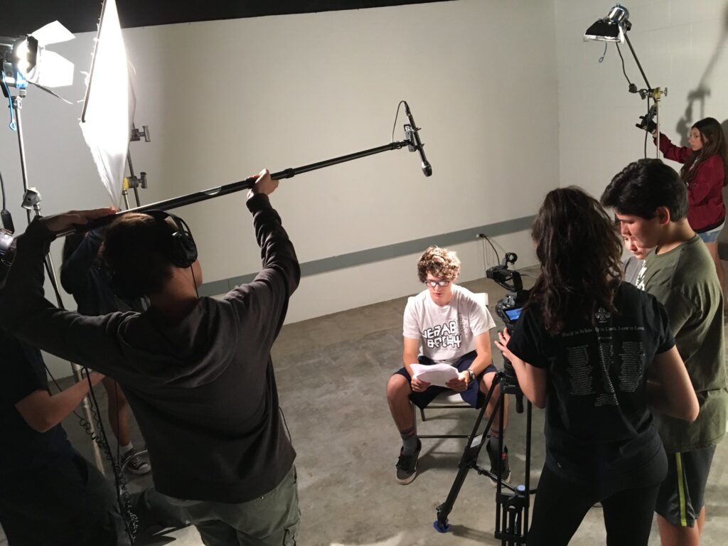 A scene showing a teen boy being interviewed, boom mics and other camera equipment visible. 