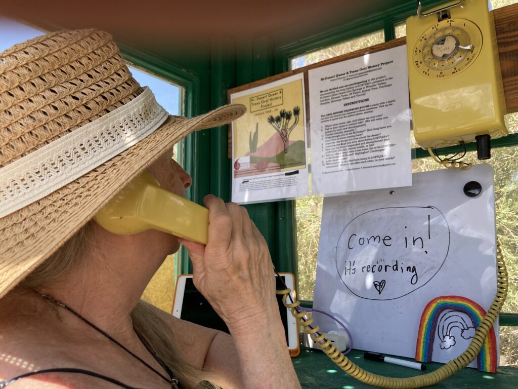 A woman wearing a straw hat holds a yellow telephone up to her ear inside a telephone booth. Words "Come in! it's recording" are on a piece of paper on the wall.