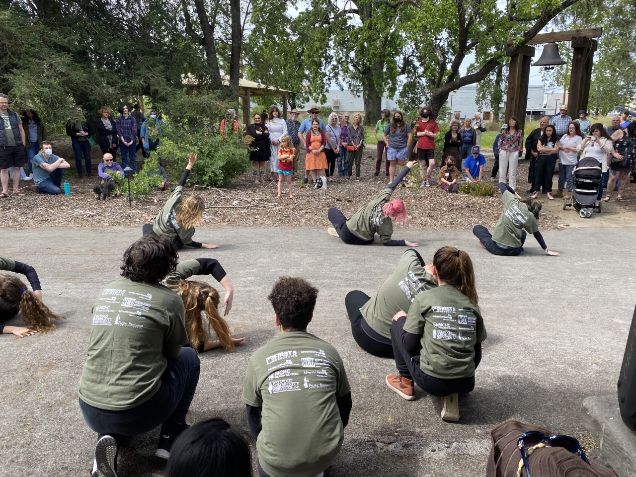 Youth dancers wearing forest green long-sleeve shirts and black pants performing for a crowd in a forested area outside.