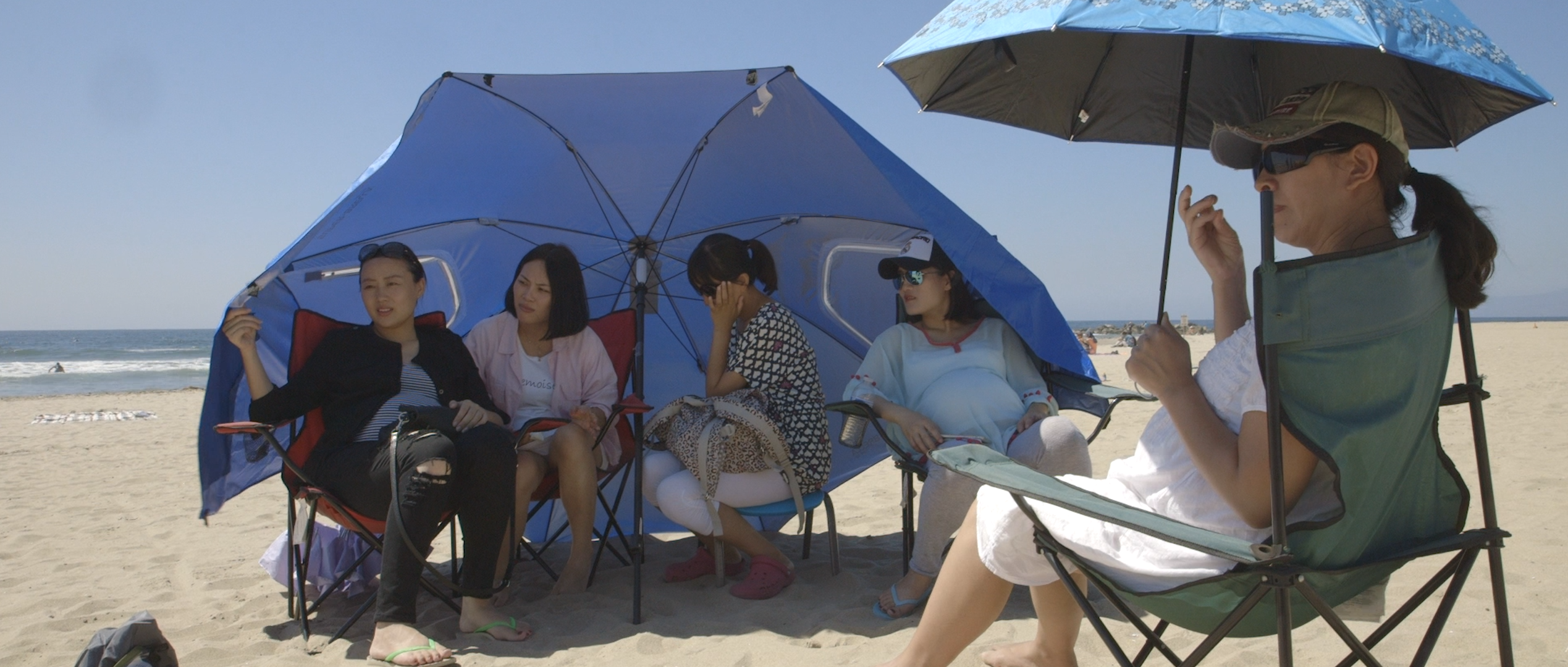 Five women sit under umbrellas in the sun on a beach.