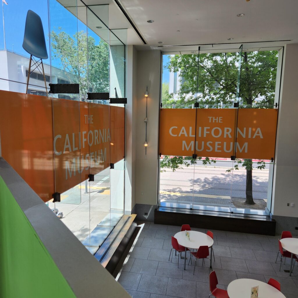 View of a large atrium with tall galss windows. Banners in the windows read The California Museum against an orange background.