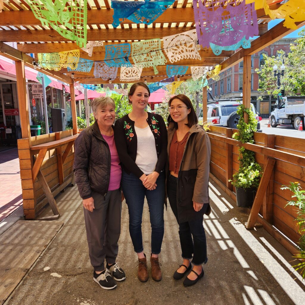 Three women stand outside and pose for the camera under a wooden canopy lined with colorful paper banners.